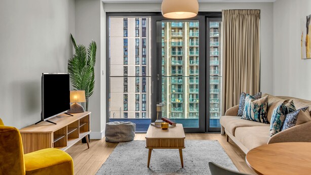 A modern living room with a sofa, a coffee table, a TV, and a large window with curtains. - Vibrant One Bed Apartment in Wembly by MySquare (Wembley Park)