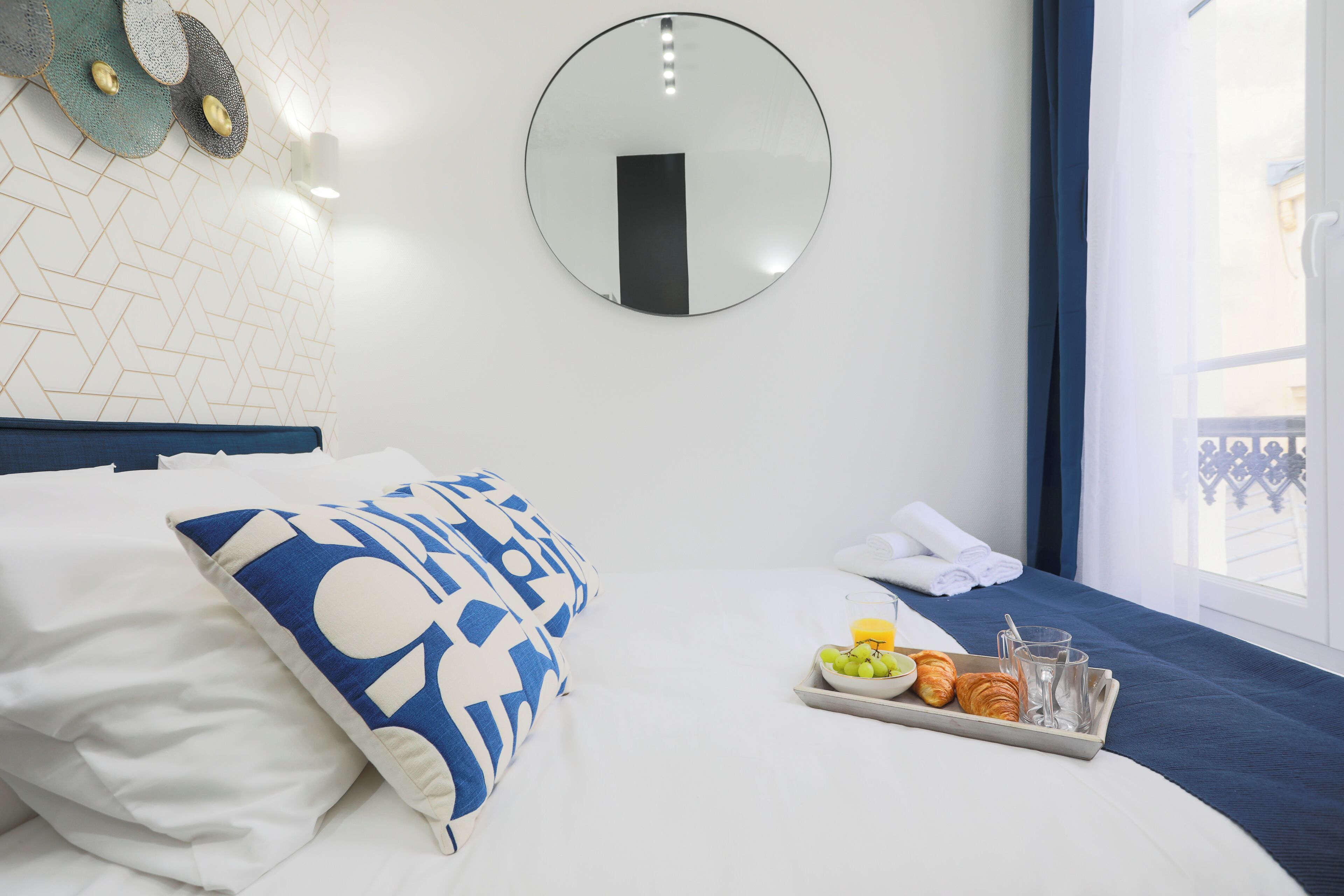Habitación de hotel con cama, bandeja con comida y bebidas, espejo redondo y ventana con vistas.