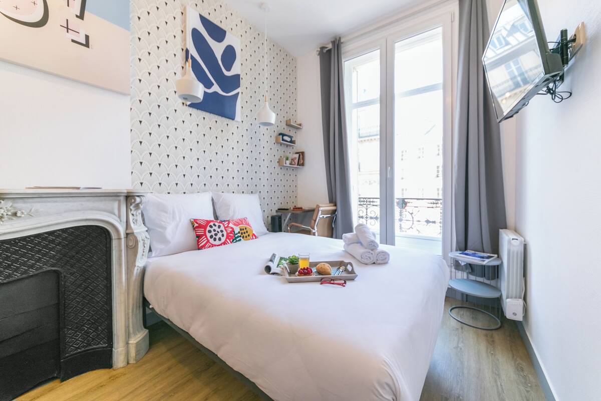 A hotel room with a bed, a small table with breakfast items, a radiator, and a window with curtains.