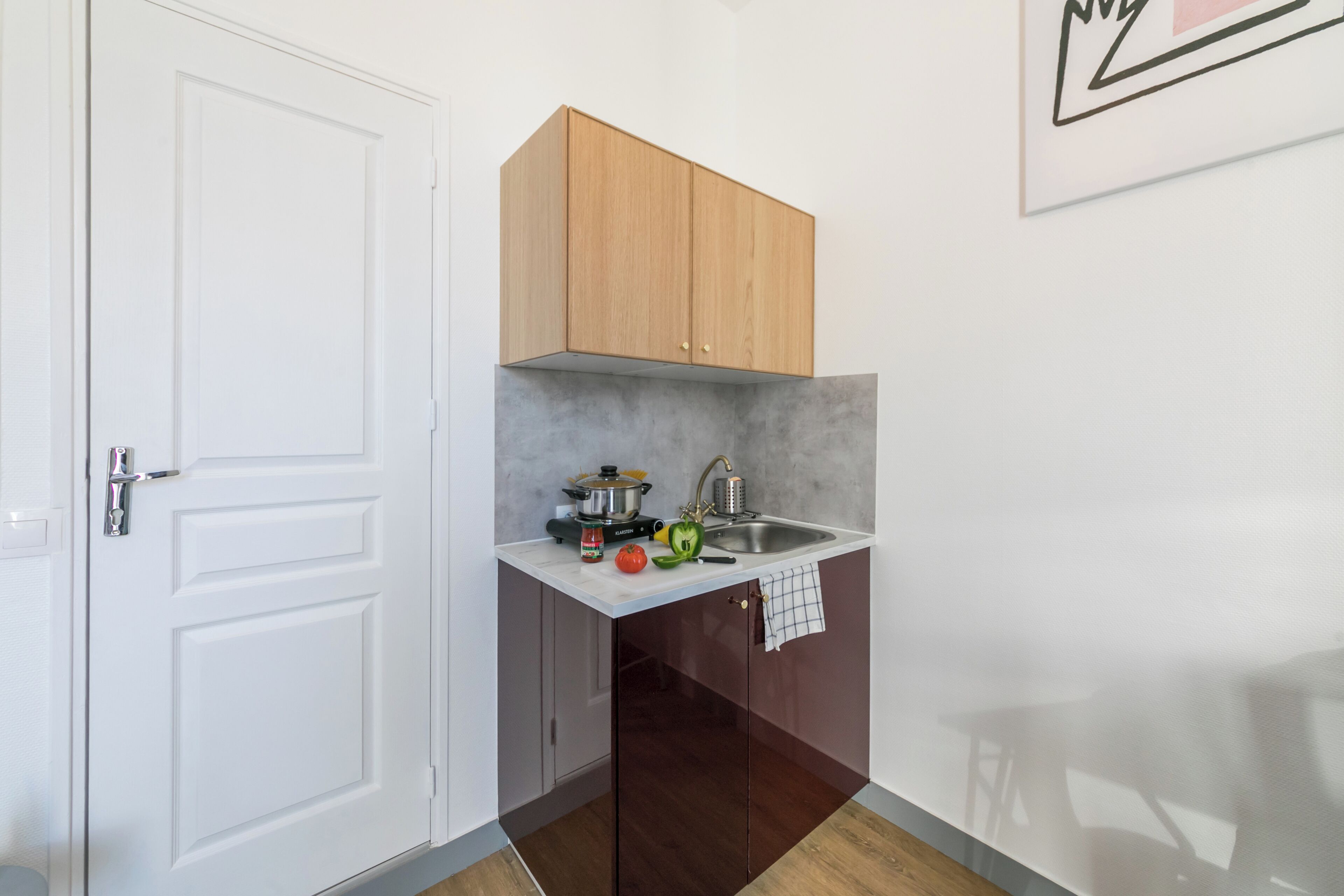 Una cocina compacta con una puerta blanca, un mueble de madera, un fregadero y una encimera con una olla, un hervidor y algunas verduras.