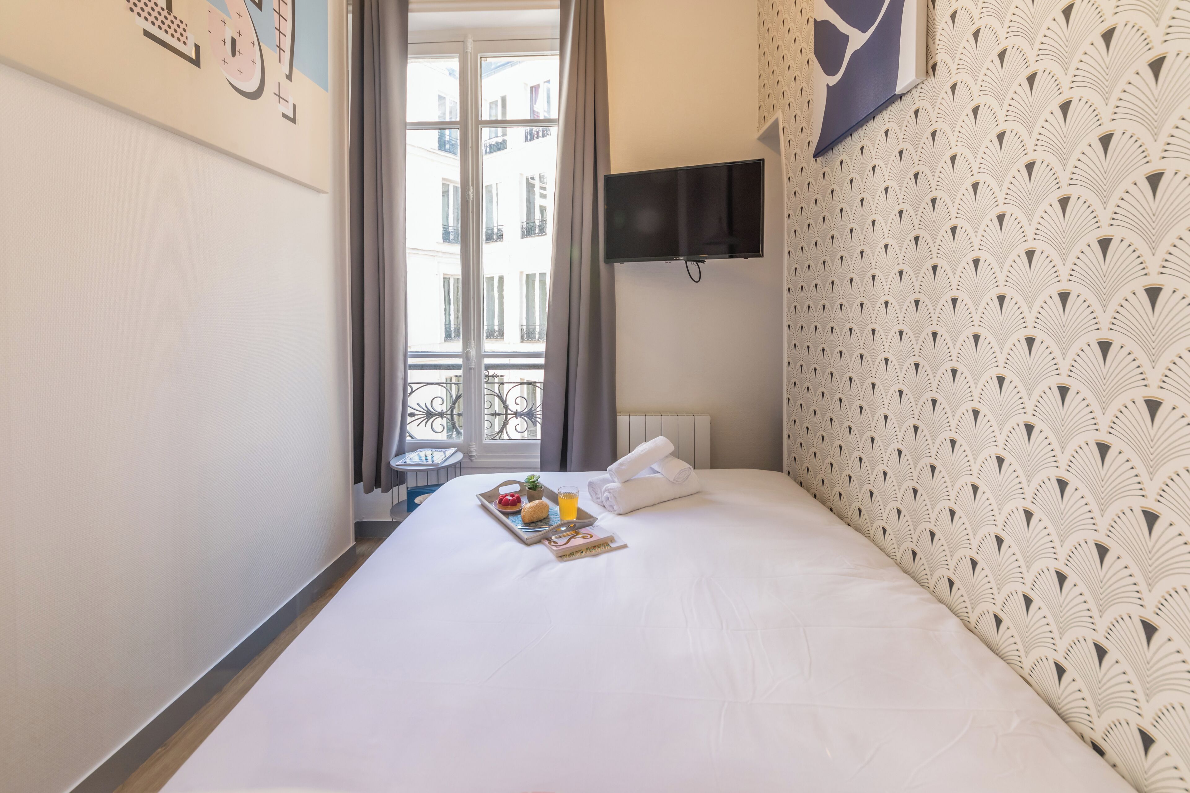 Habitación de hotel con cama, televisión montada en la pared, ventana con cortinas y balcón con barandilla.