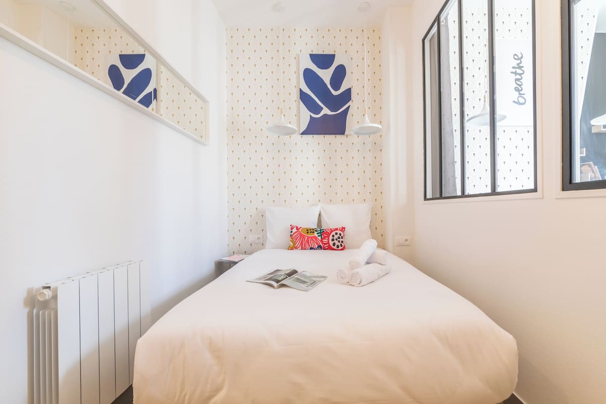 A neatly made bed with a white comforter, a colorful pillow, and a rolled towel. There is a magazine on the bed, and a window with a view of the outside.