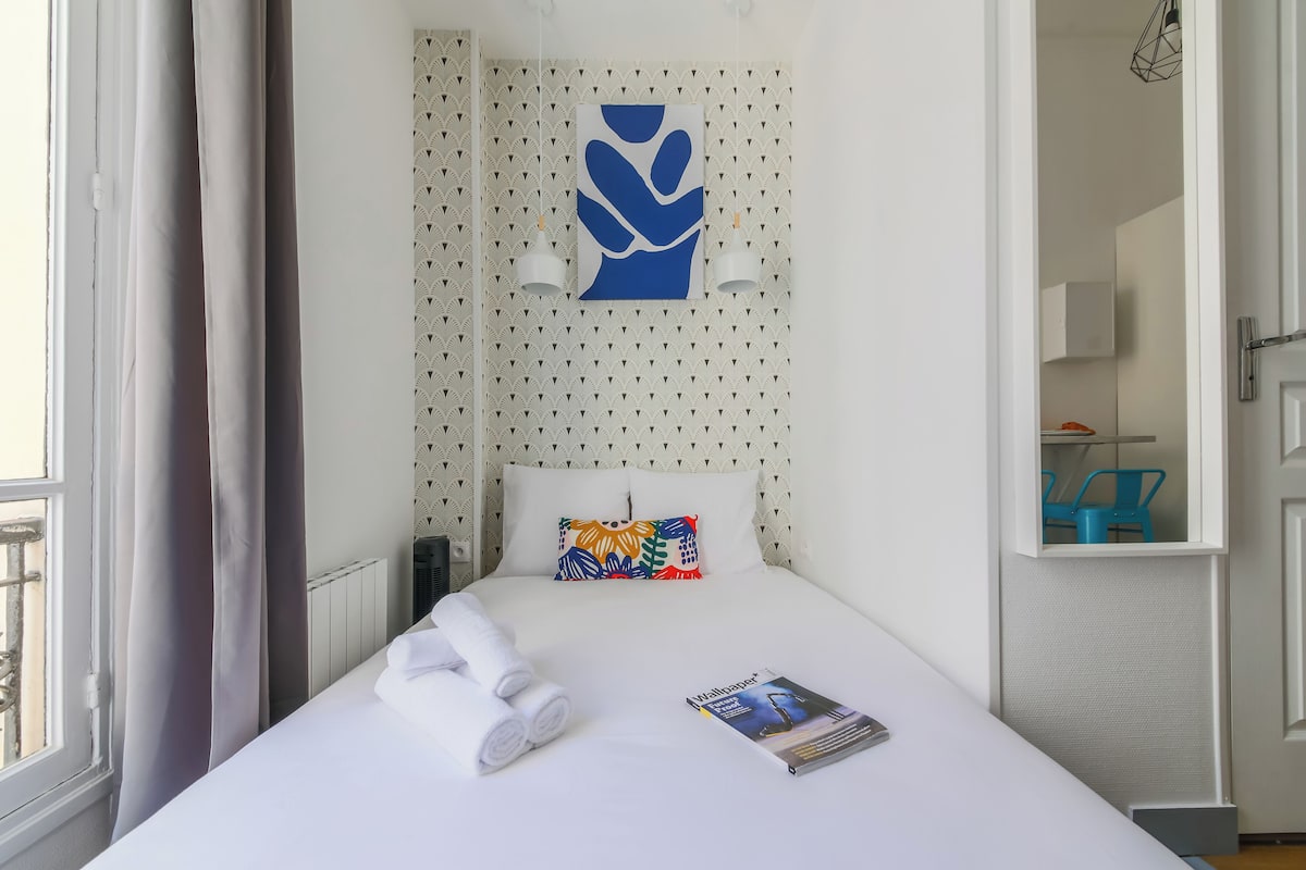A hotel room with a neatly made bed, a colorful pillow, a magazine on the bed, and a window with curtains.