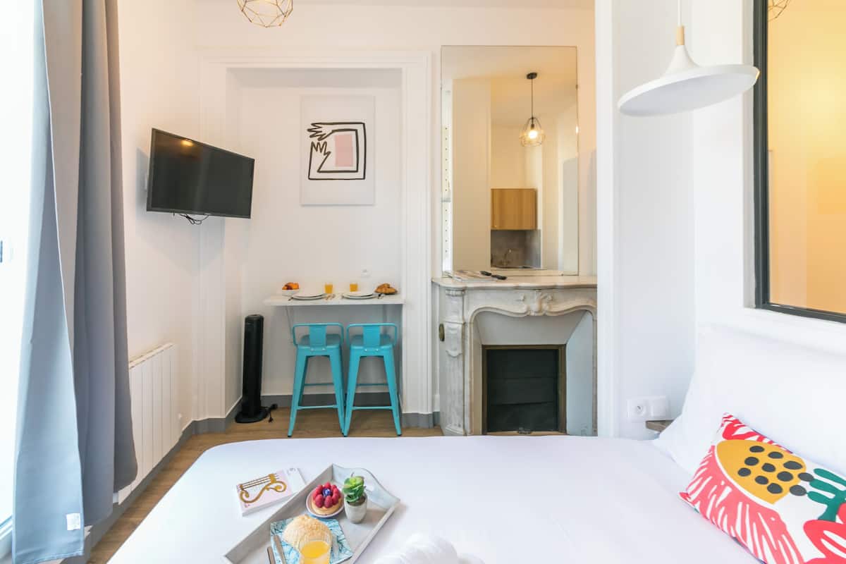 A bedroom with a bed, a TV mounted on the wall, a small table with chairs, and a fireplace.