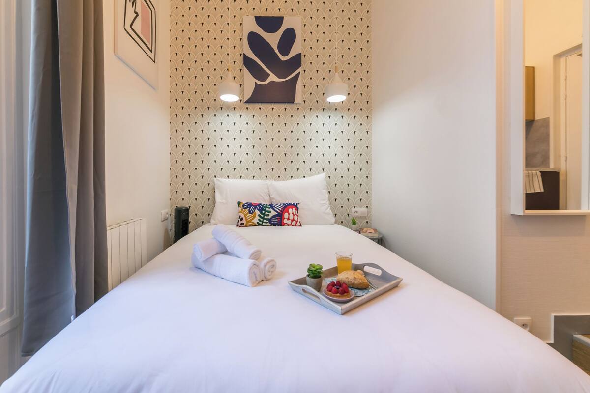 A neatly made bed with white linens, a tray with breakfast items, and a colorful pillow.