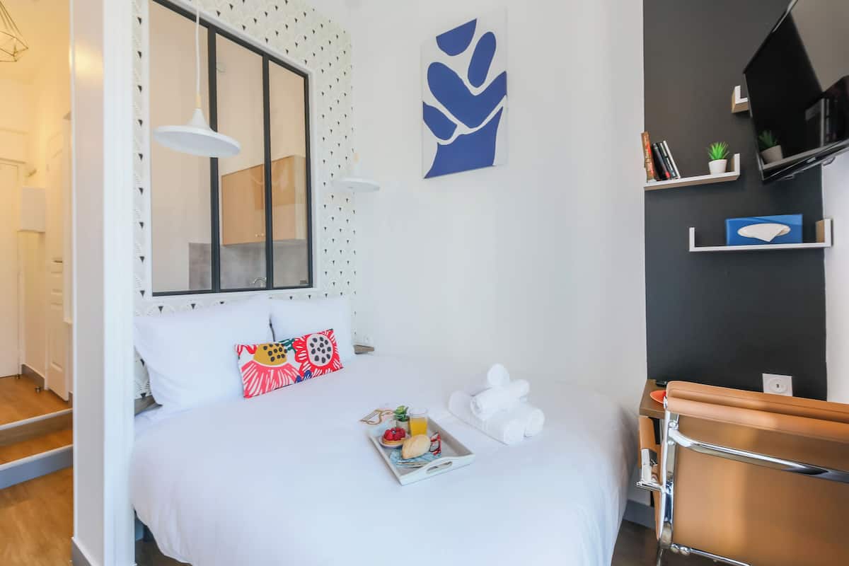 A modern bedroom with a bed, a bedside table with a tray of breakfast, a mirror, a wall art piece, and a TV mounted on the wall.