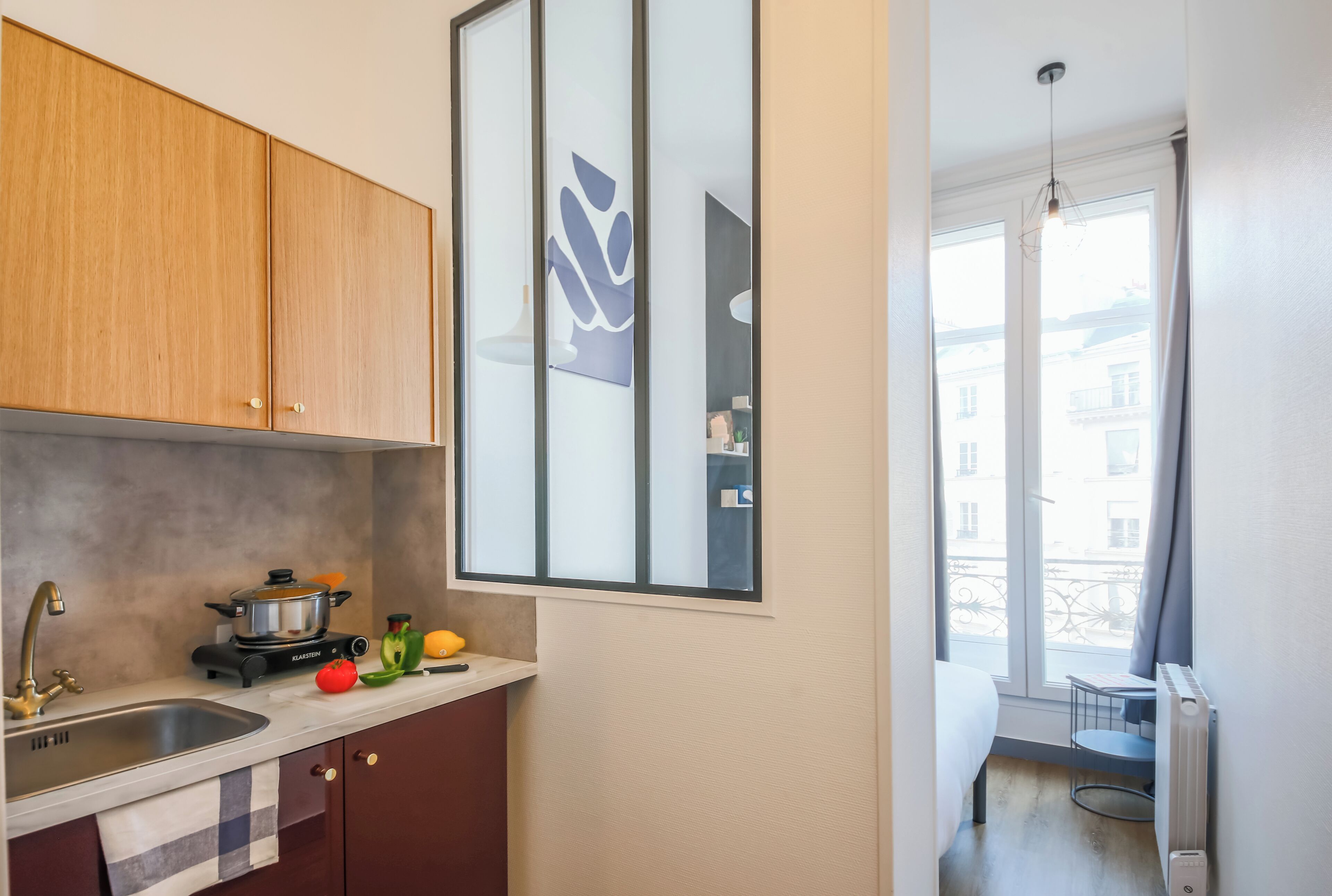 Une cuisine compacte équipée d’une cuisinière, d’un évier et de meubles en bois. Une fenêtre offre une vue sur les bâtiments environnants.