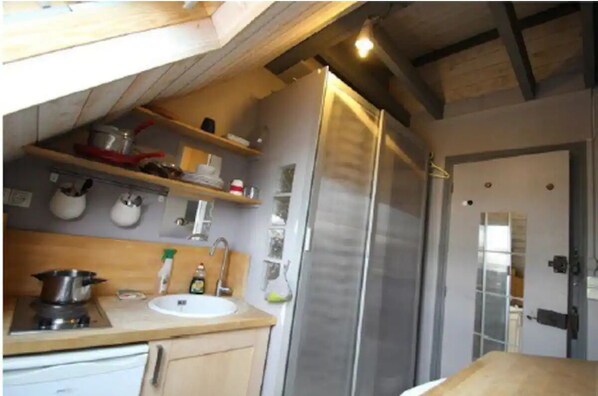 Compact kitchen with a refrigerator, sink, and wooden countertop, featuring exposed beams and a door to the adjacent room.