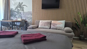 A bedroom with a bed, a TV mounted on the wall, a desk with chairs, and a view of palm trees through a window. - TUDOR STUDIO (Kaprun)