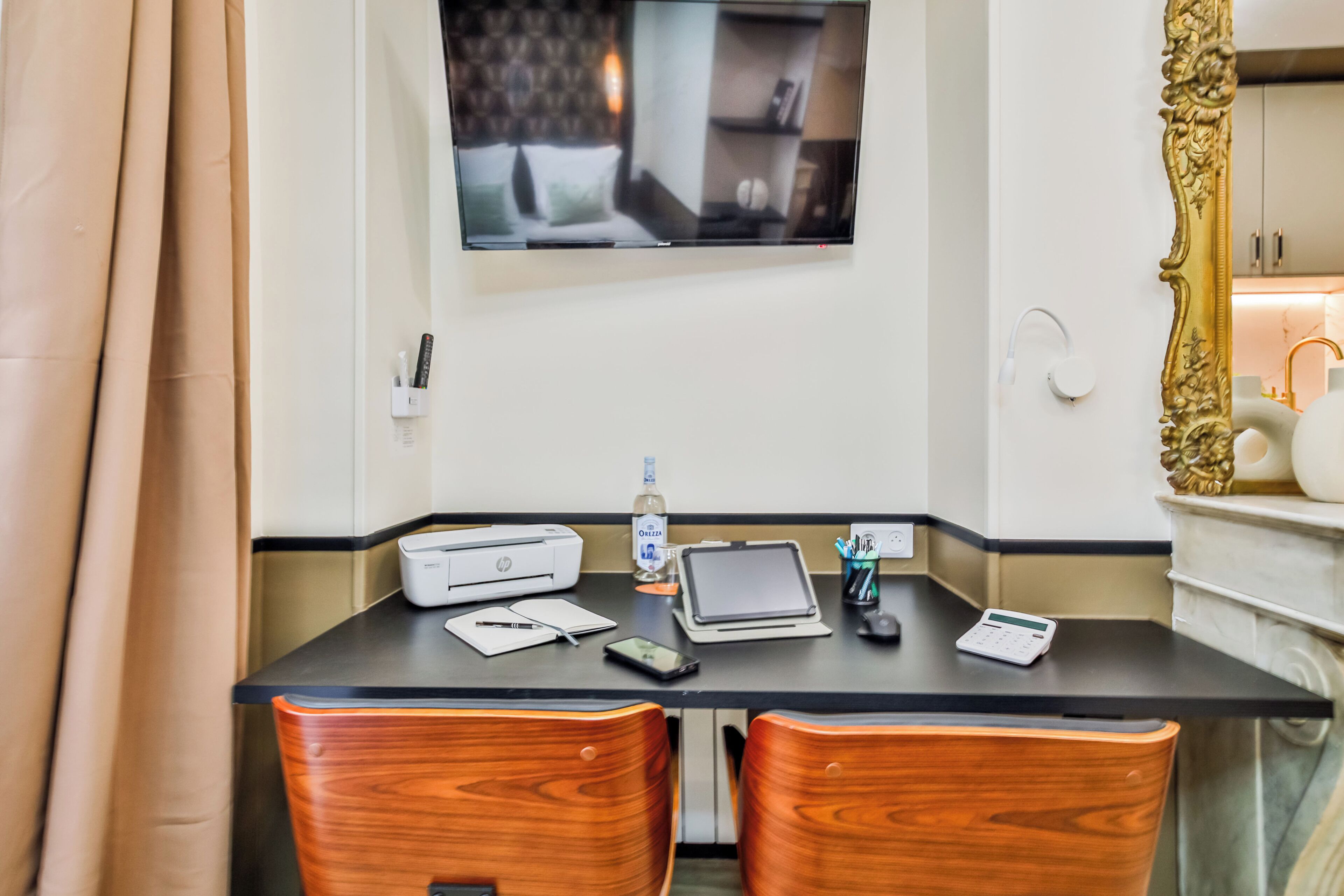 Ein Hotelzimmer mit einem an der Wand befestigten Flachbildfernseher, einem Schreibtisch mit Drucker, Tablet, Smartphone und Taschenrechner. Am Schreibtisch stehen zwei Holzst ühle.