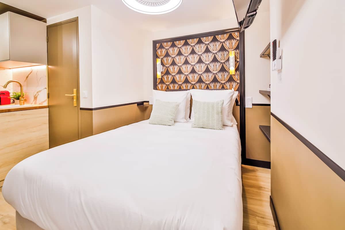A modern hotel room with a large bed, a small bedside table, a built-in shelf, and a bathroom with a toilet and sink.