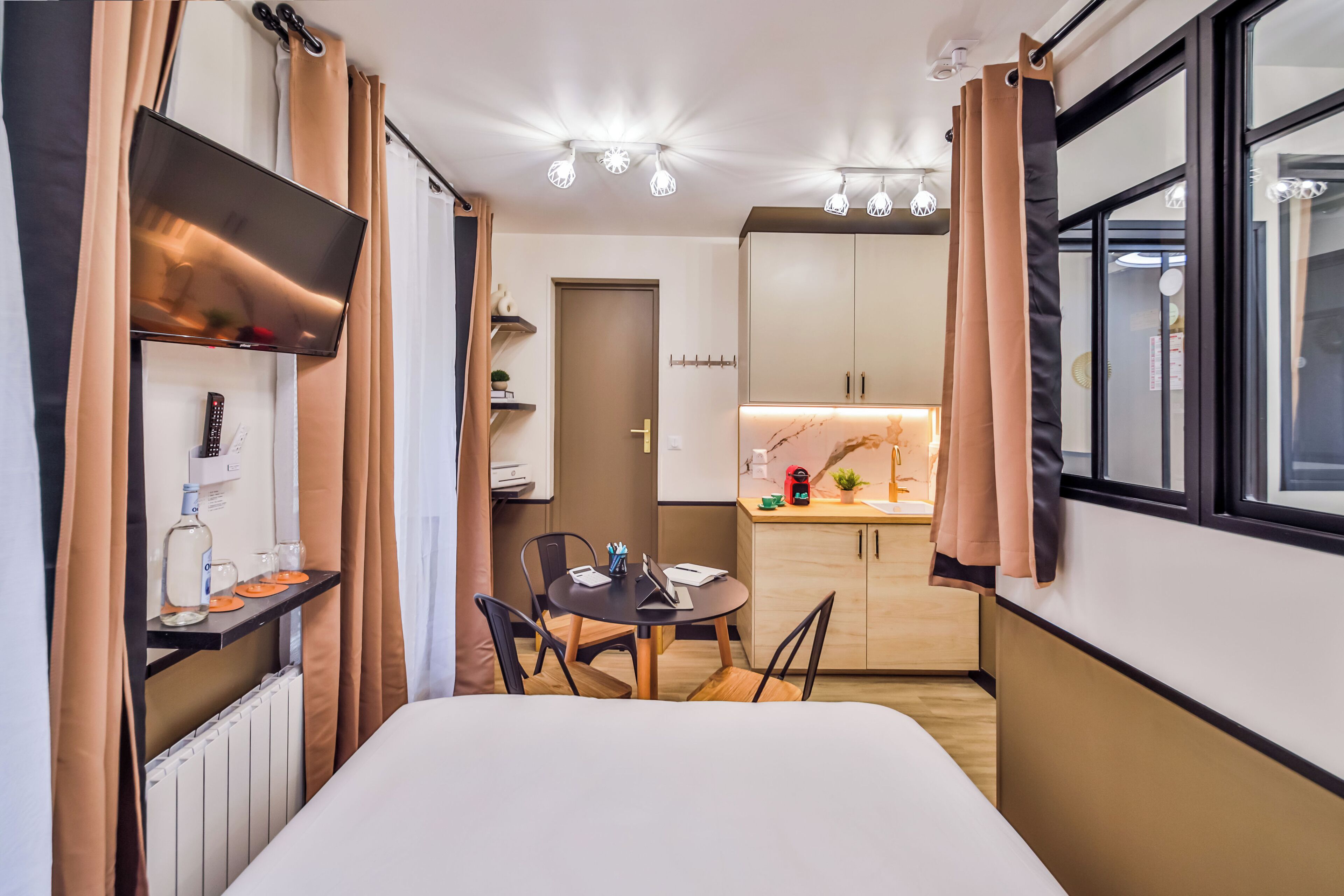 Una habitación de hotel compacta con una cama, una pequeña mesa de comedor con sillas, una kitchenette y un ventanal grande.