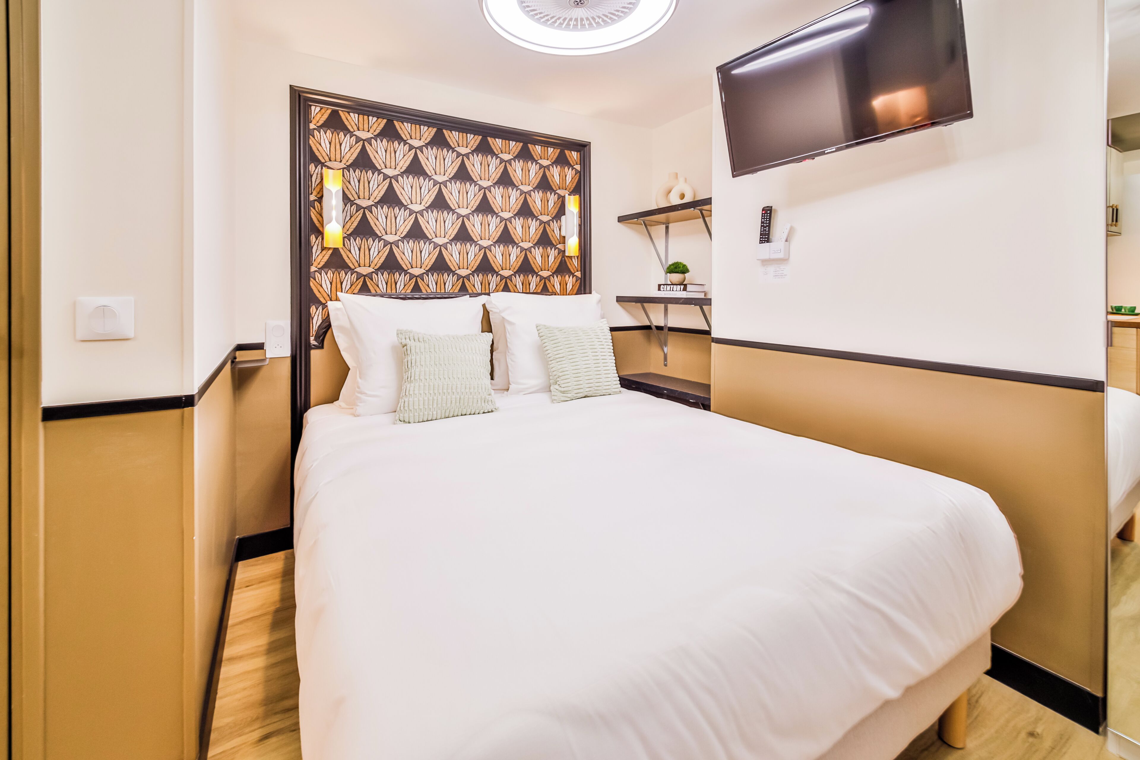A hotel room with a large bed, a TV mounted on the wall, a small desk with a plant, and a decorative headboard.