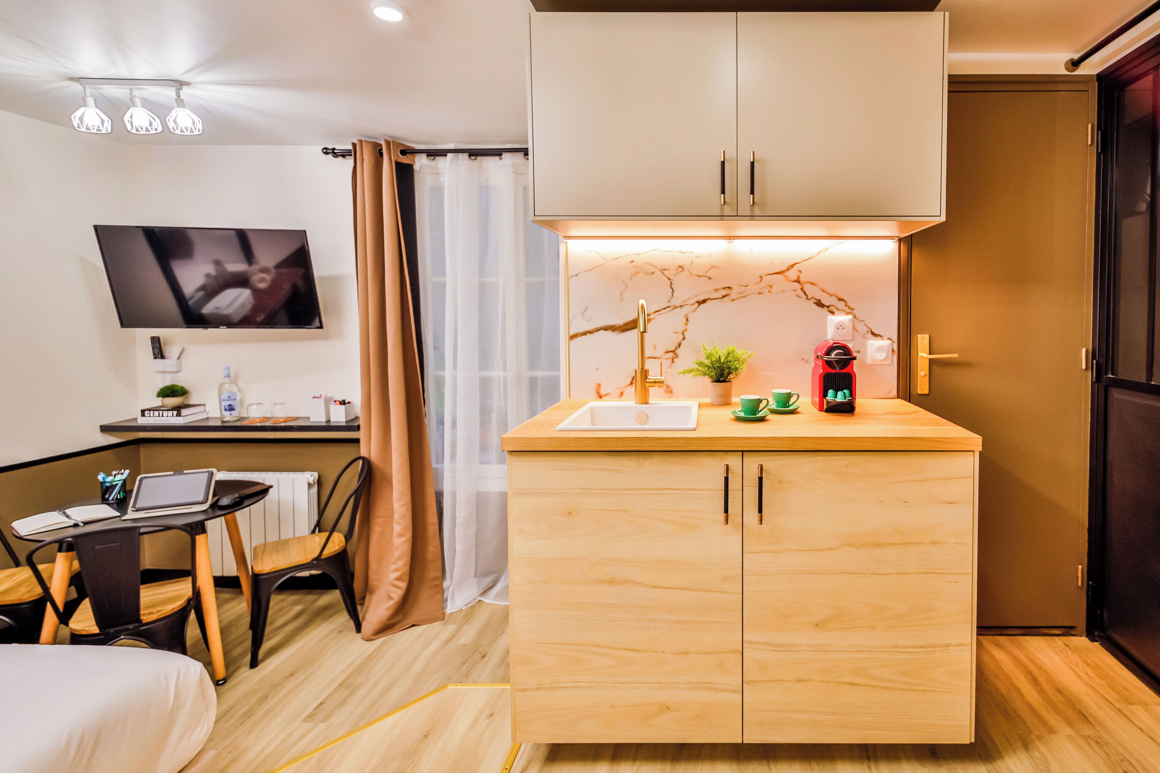 Een compacte keuken met een houten aanrecht, een spoelbak en een klein plantje. Er is een eethoek met een tafel en stoelen, en een televisie aan de muur bevestigd.