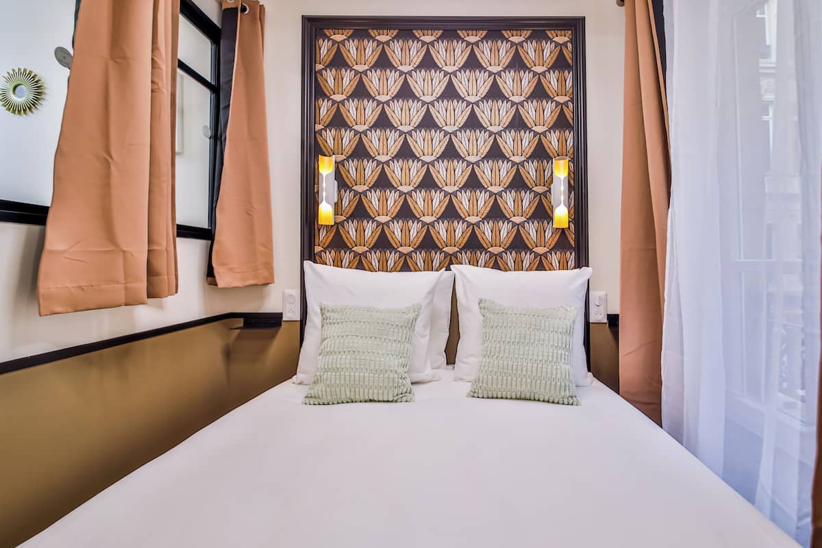 A hotel room with a bed, two pillows, a patterned headboard, and curtains.