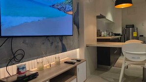 Un soggiorno moderno con un'ampia televisione a schermo piatto montata a parete, un mobile basso in legno, un tavolo da pranzo bianco con sedie e un'area cucina sullo sfondo.
