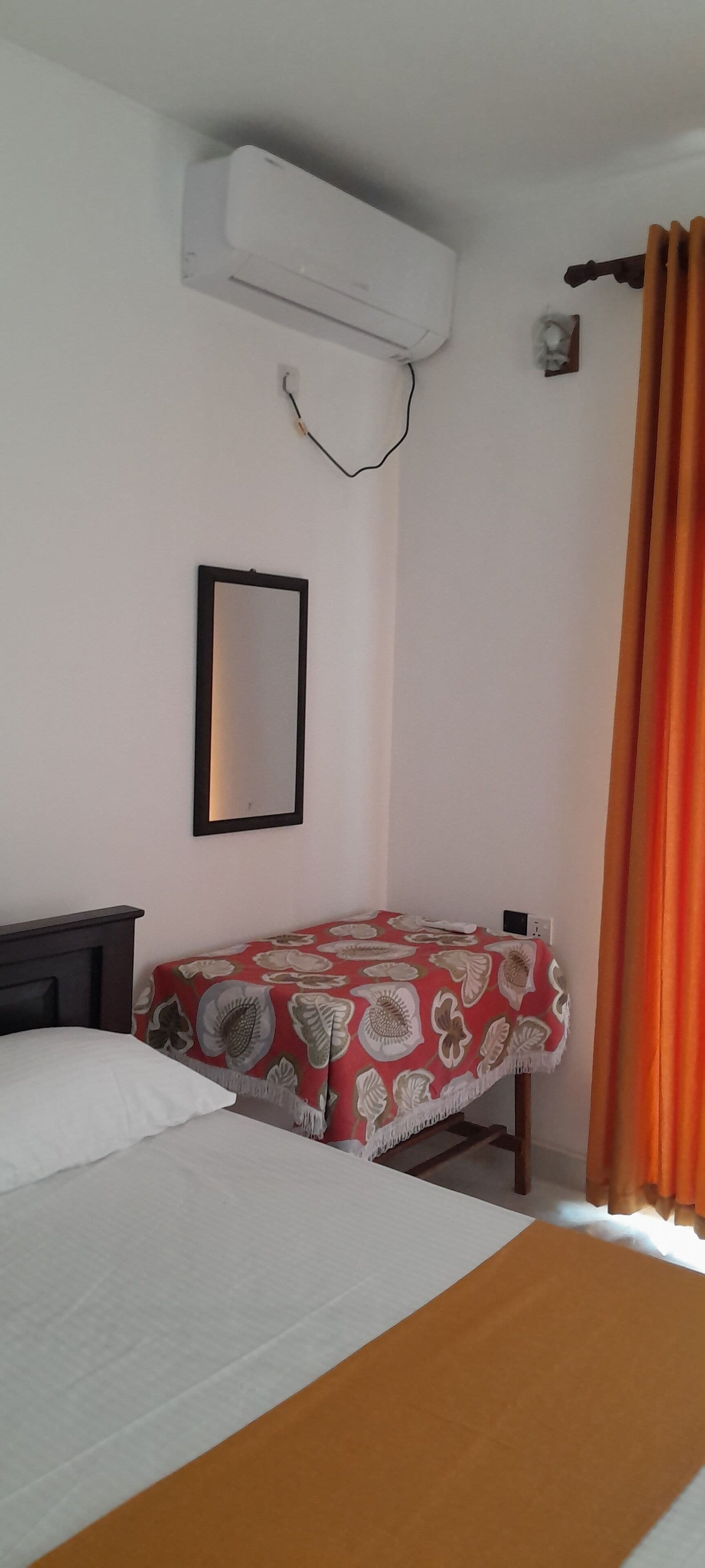 A bedroom with a bed, a bedside table, an air conditioning unit, a mirror, and a window with curtains.