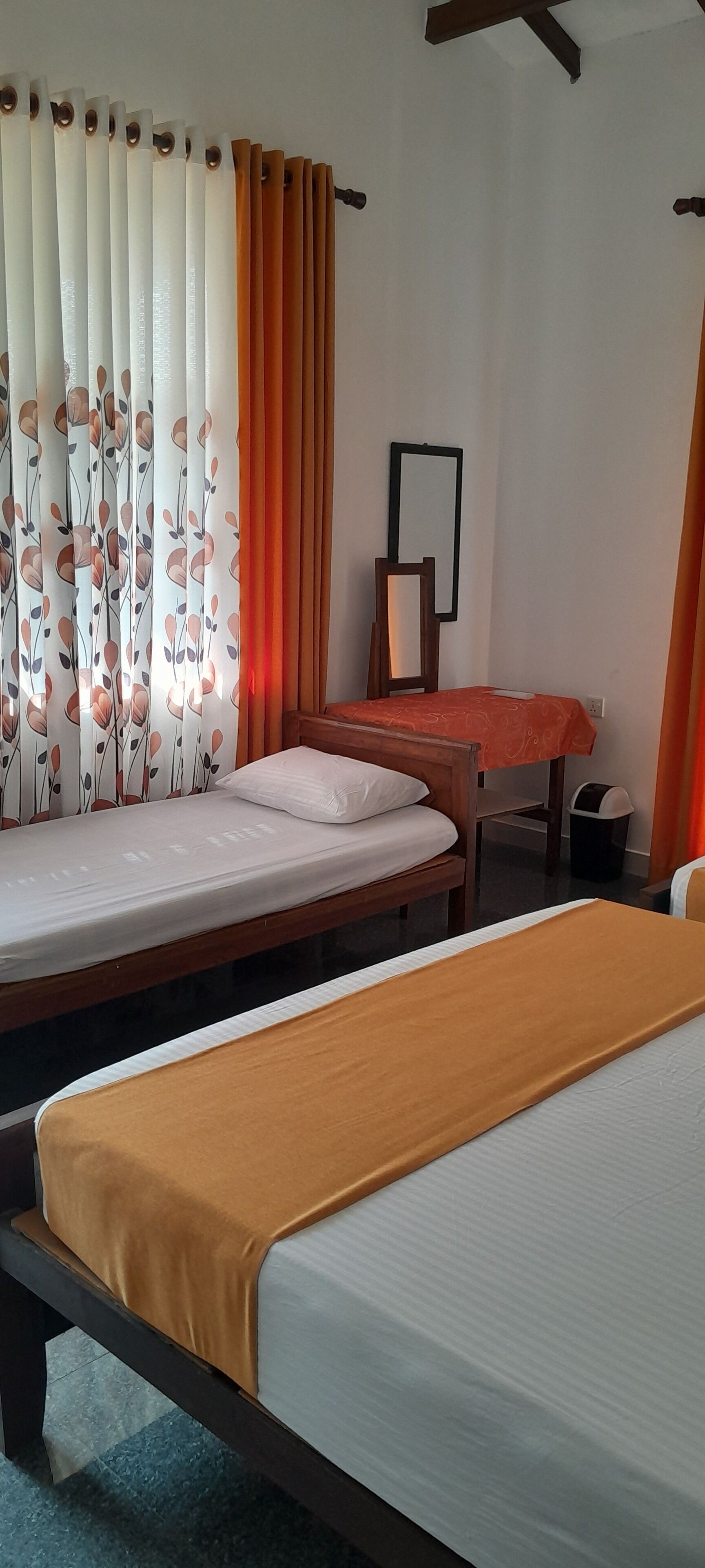 A room with two beds, one with a brown bedspread and the other with a white bedspread. There is a wooden chair and a small table with a red tablecloth. The room has white walls and a window with orange curtains.