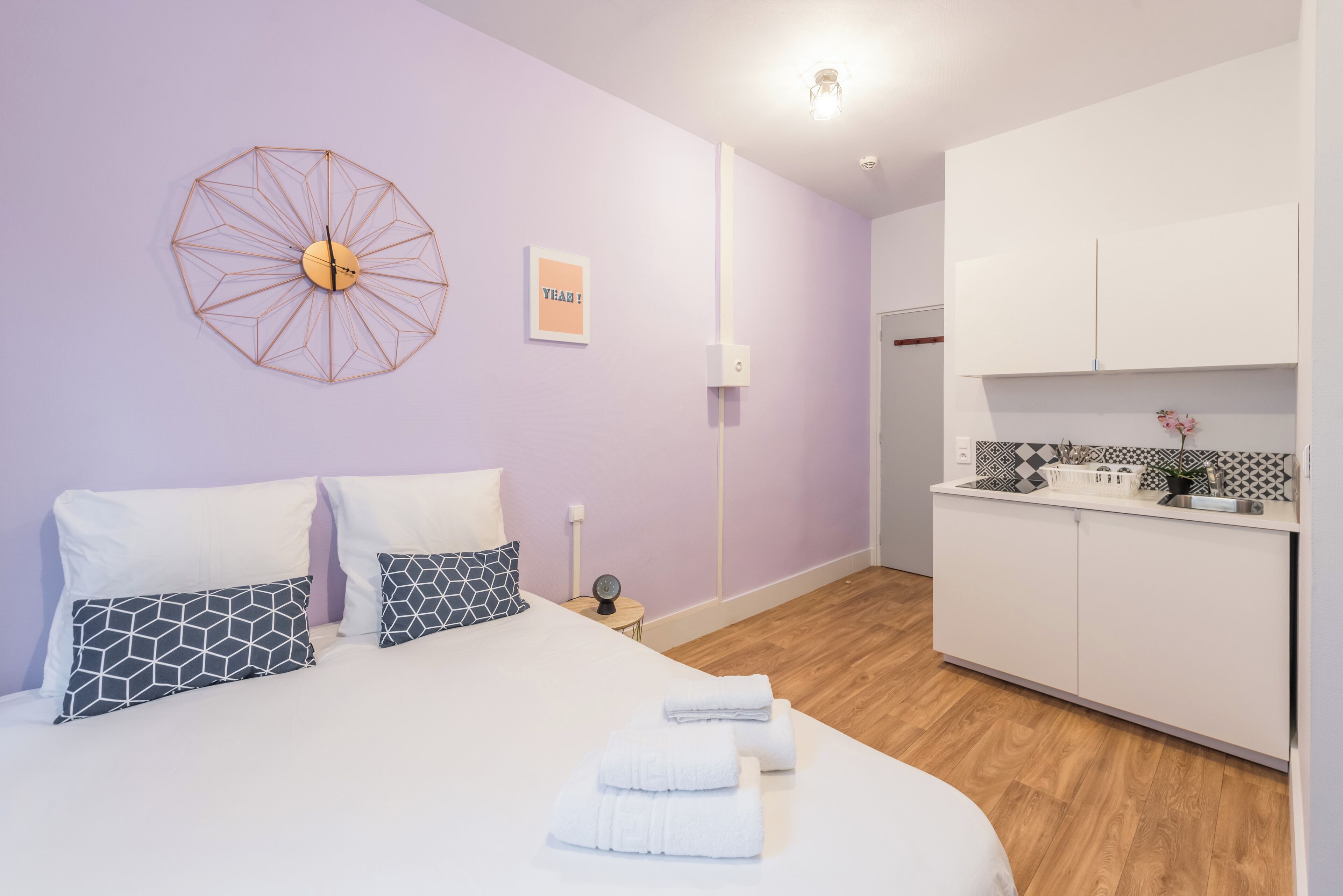 A modern bedroom with a large bed, geometric-patterned pillows, a wall clock, and a small bedside table.