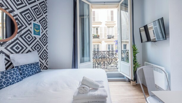 A hotel room with a bed, a TV, a balcony with a view of buildings, and a patterned wall. - WS Hôtel de Ville - Musée Pompidou (Paris)