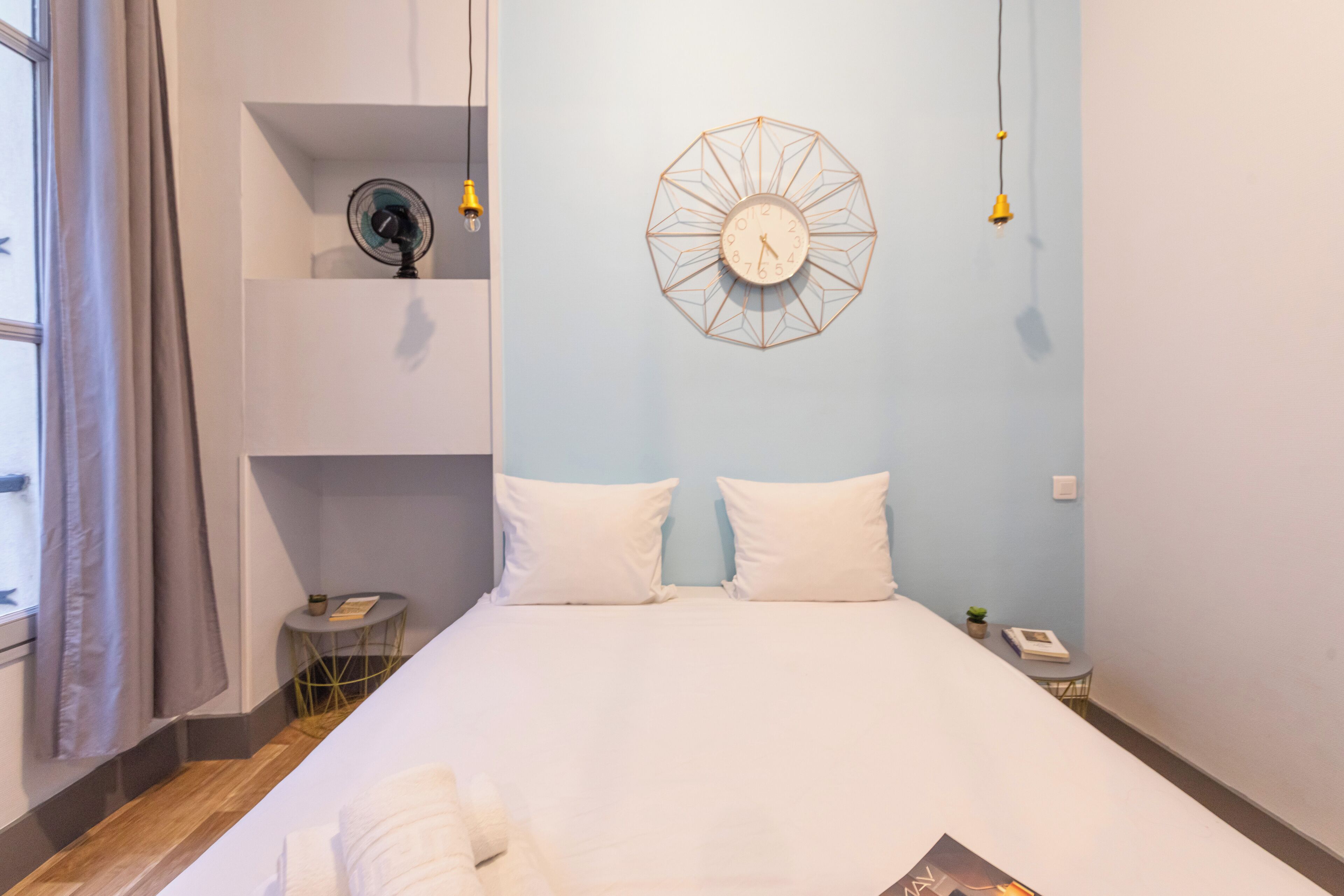 Uma cama bem arrumada com roupa de cama branca, um relógio fixado na parede e uma mesinha com um ventilador e um abajur.