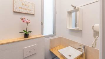A modern bathroom with a wooden shelf, a white sink, a mirror, a hairdryer, and a toilet paper roll.