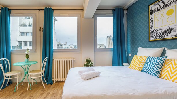 A hotel room with a bed, a small table with chairs, a view of the city through large windows, and a decorative wall piece. - WS Champs Elysées - Ponthieu (Paris)