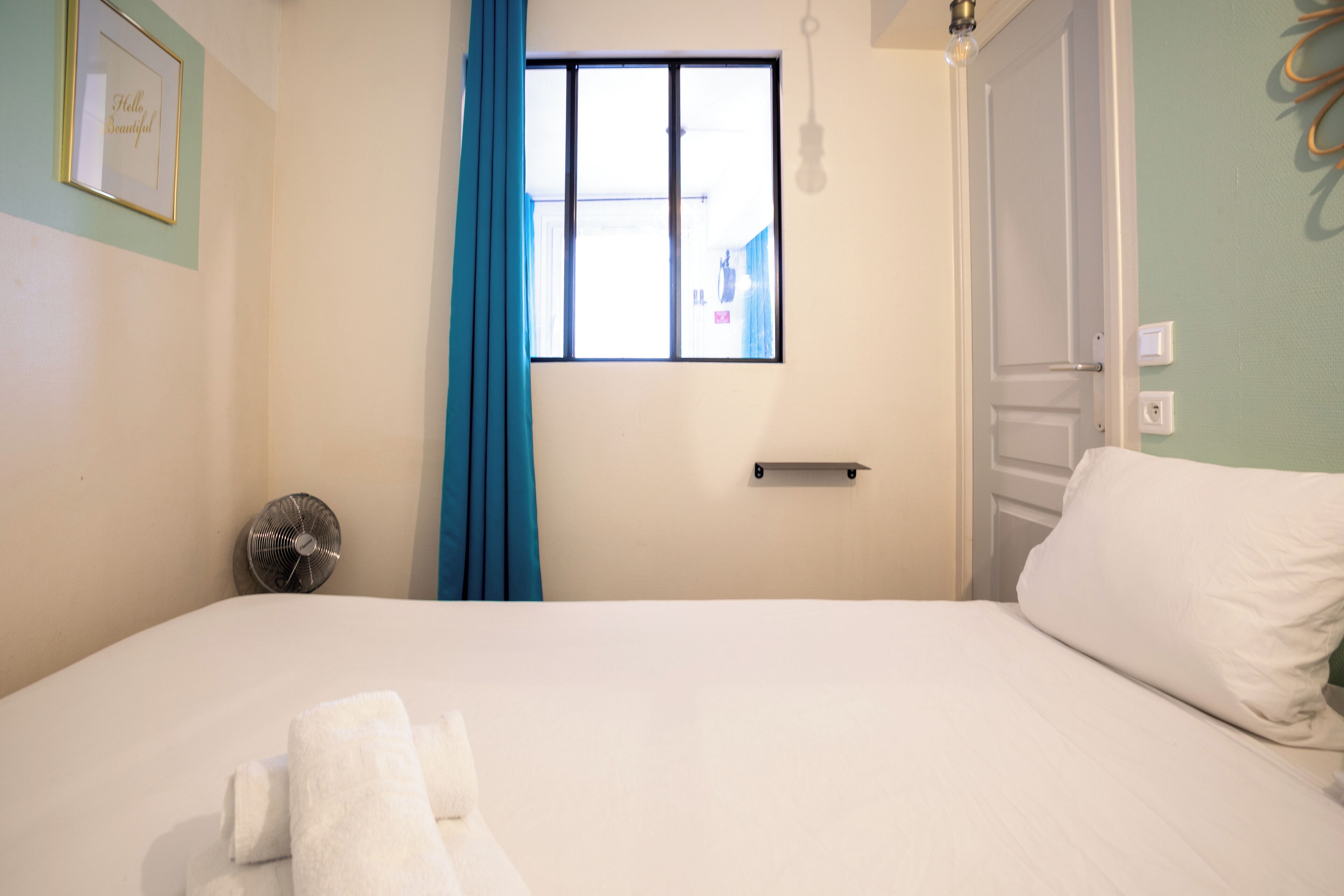 Una cama bien hecha con sábanas blancas, una cortina azul, un ventilador de pared y una ventana con vistas al exterior.