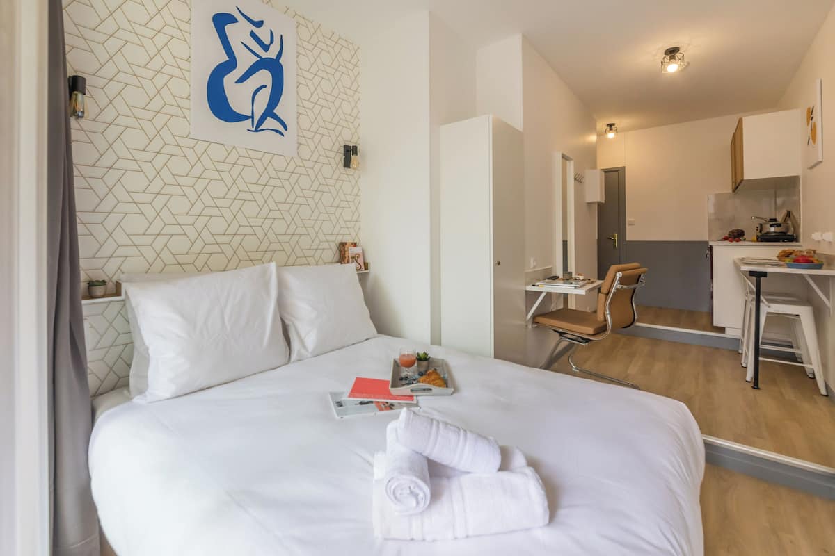Quarto de hotel com cama bem arrumada, escrivaninha com cadeira e uma kitchenette ao fundo.