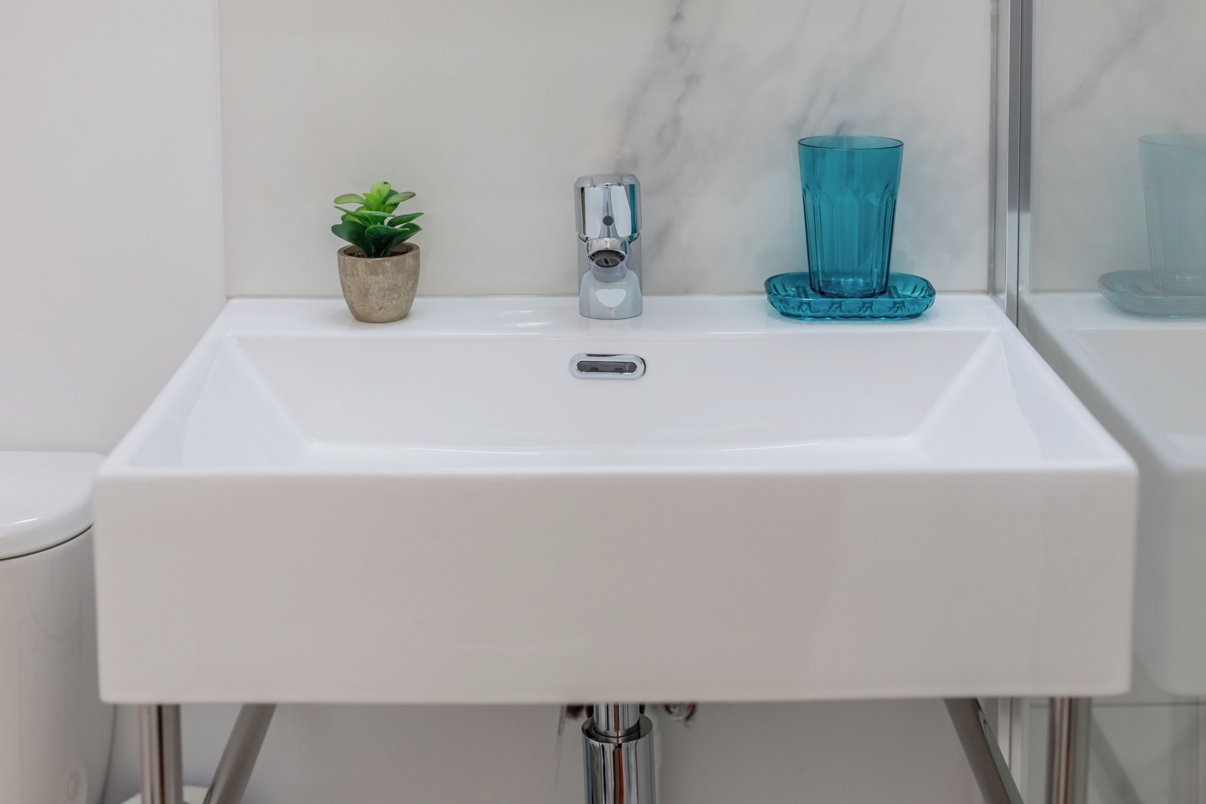 Ein weißes Waschbecken mit modernem Wasserhahn, eine kleine Topfpflanze und ein blaues Glas auf der Ablagefläche.