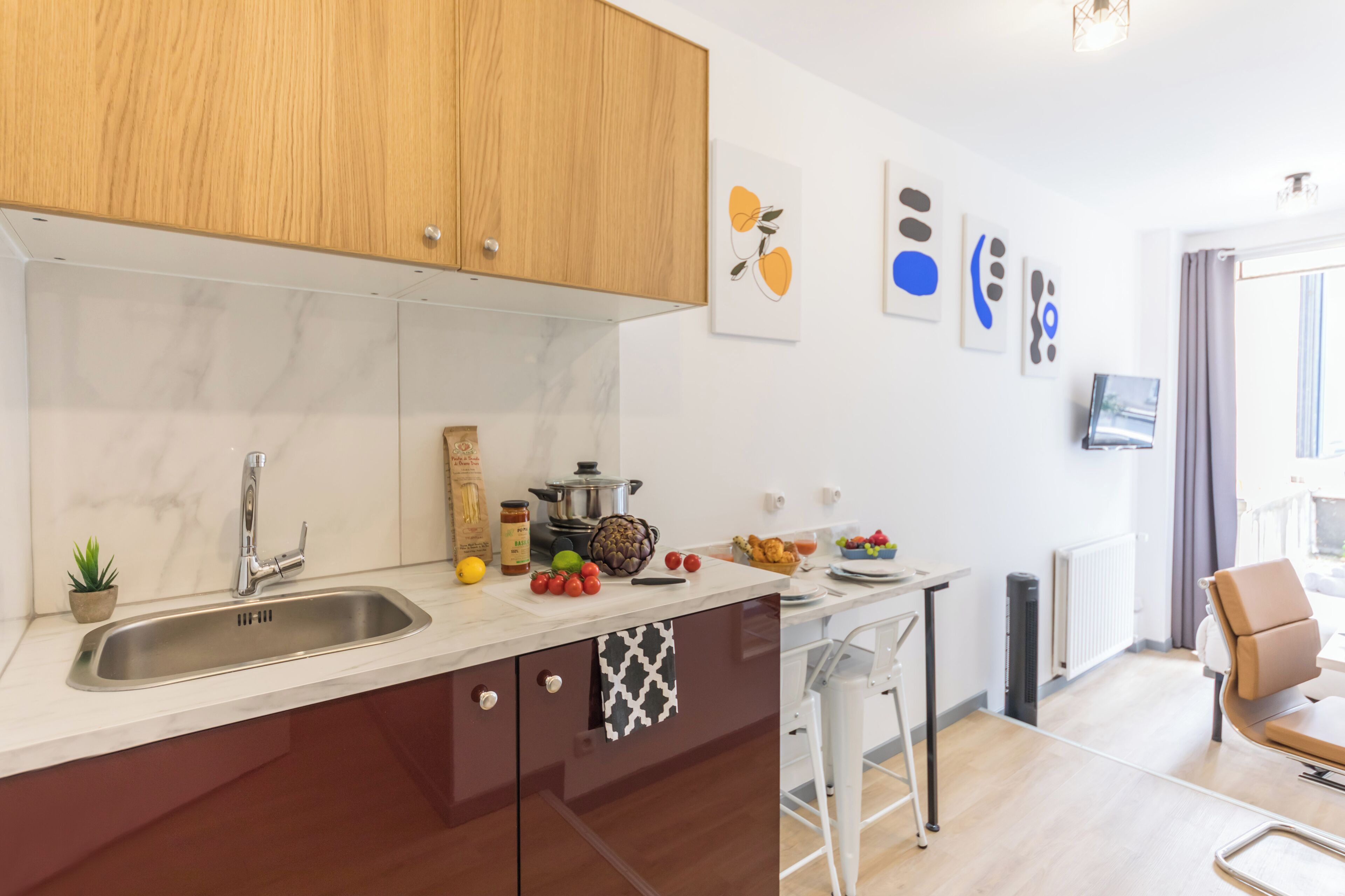A modern kitchen with a marble countertop, wooden cabinets, and a built-in stove. There is a small plant on the counter and a window with curtains.