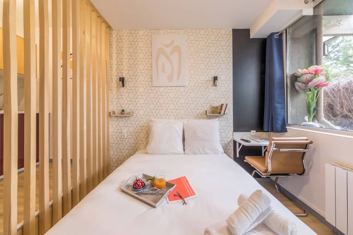 Quarto de hotel moderno com cama, uma pequena escrivaninha, uma cadeira e janela com cortinas.