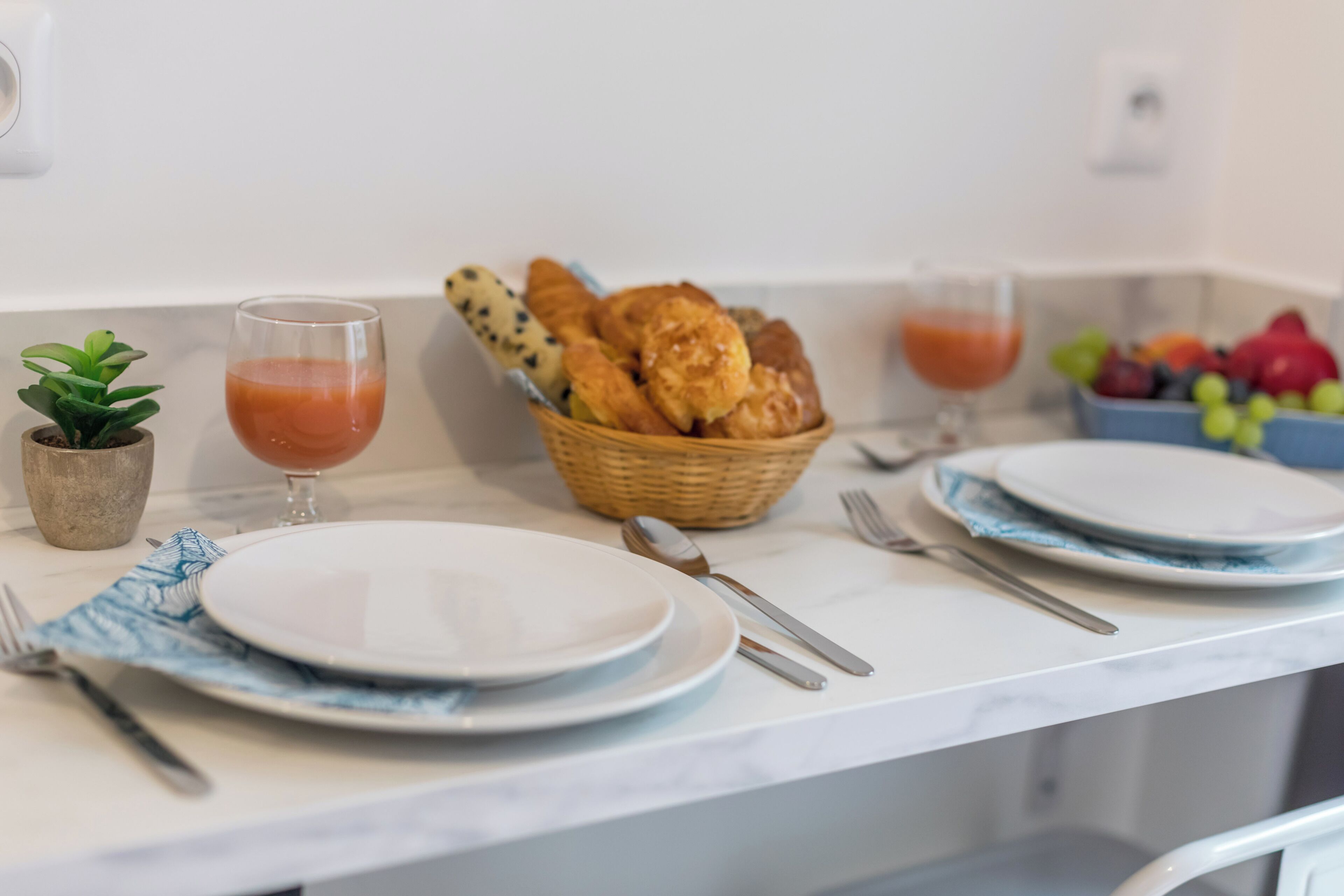 Ein Frühstückstisch mit Tellern, Besteck, einem Gebäckkorb und einer Obstschale.