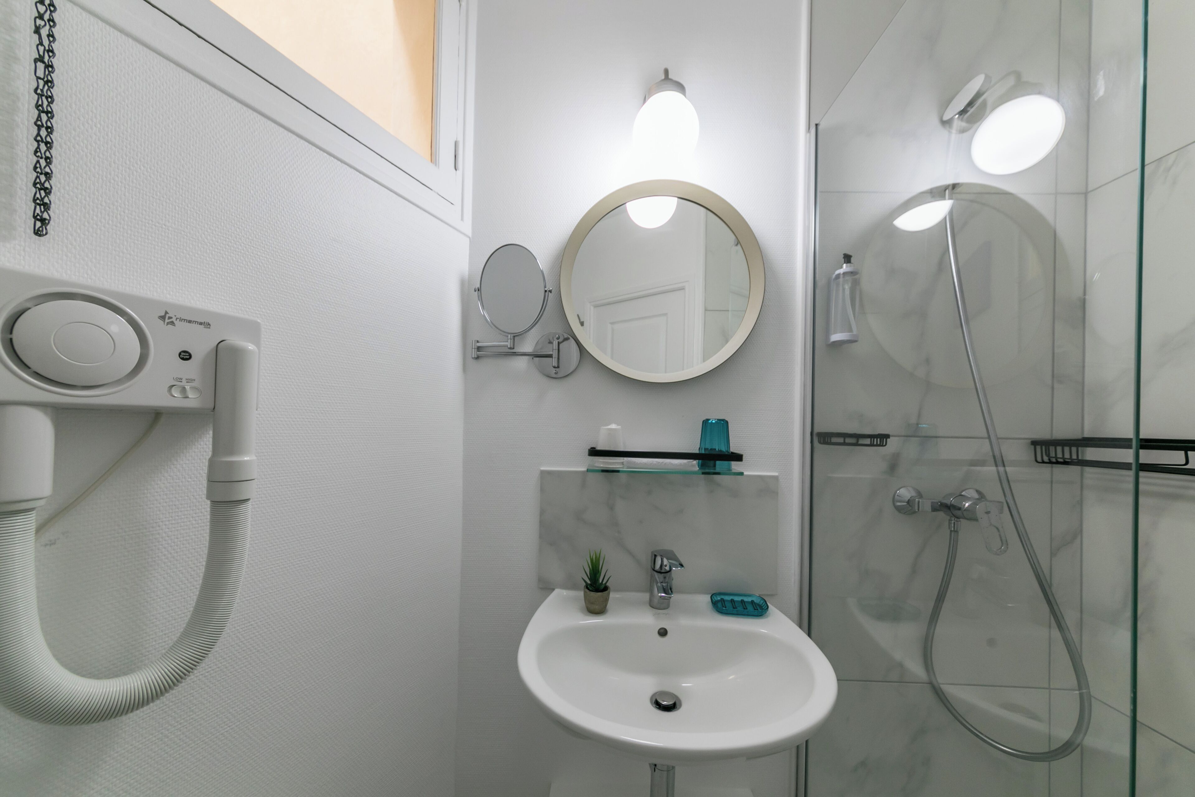 A modern bathroom with a white sink, a round mirror, a handheld mirror, a glass shower enclosure, and a handheld showerhead.