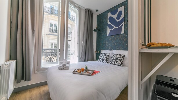 A hotel room with a large bed, a window with curtains, a radiator, a wall art piece, and a tray with breakfast items. - WS Opéra - Rossini (Paris)