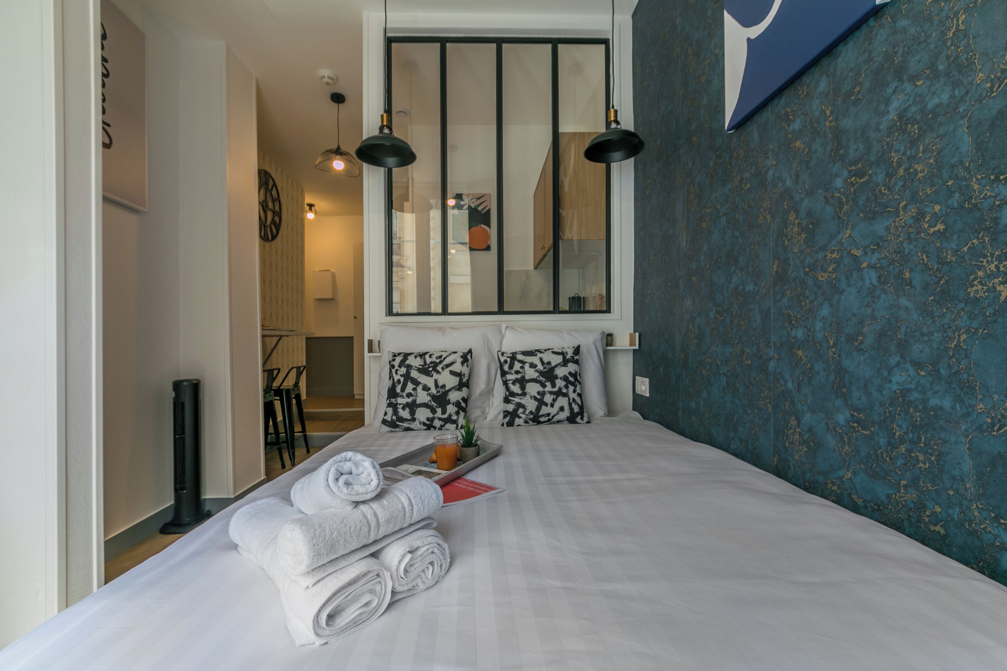 Habitación de hotel con cama, toallas, una bandeja con una bebida y vista a la cocina por una ventana.