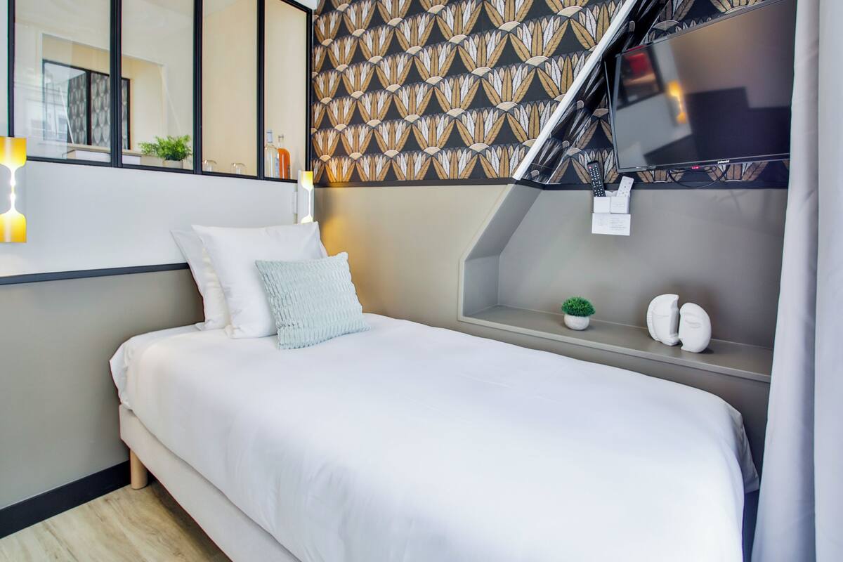 A modern hotel room with a neatly made bed, a small bedside table with a plant, and a large window with a view of the city.