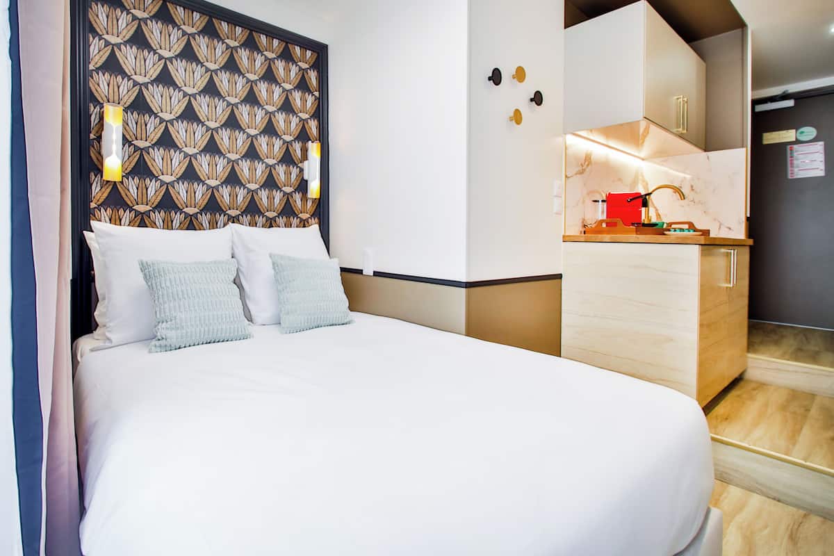A hotel room with a large bed, a wooden headboard, a small kitchenette, and a wooden floor.