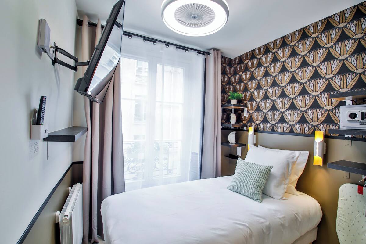 A hotel room with a bed, a TV mounted on the wall, a radiator, a window with curtains, and a patterned wallpaper.