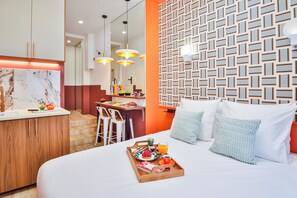A modern hotel room with a bed, a wooden side table with breakfast items, a kitchen area with a dining table, and a patterned wall. - WS Opéra - Châteaudun (Paris)