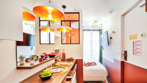 Una cocina moderna con encimera de madera, lámparas colgantes naranjas y una zona de comedor con una cama al fondo.
