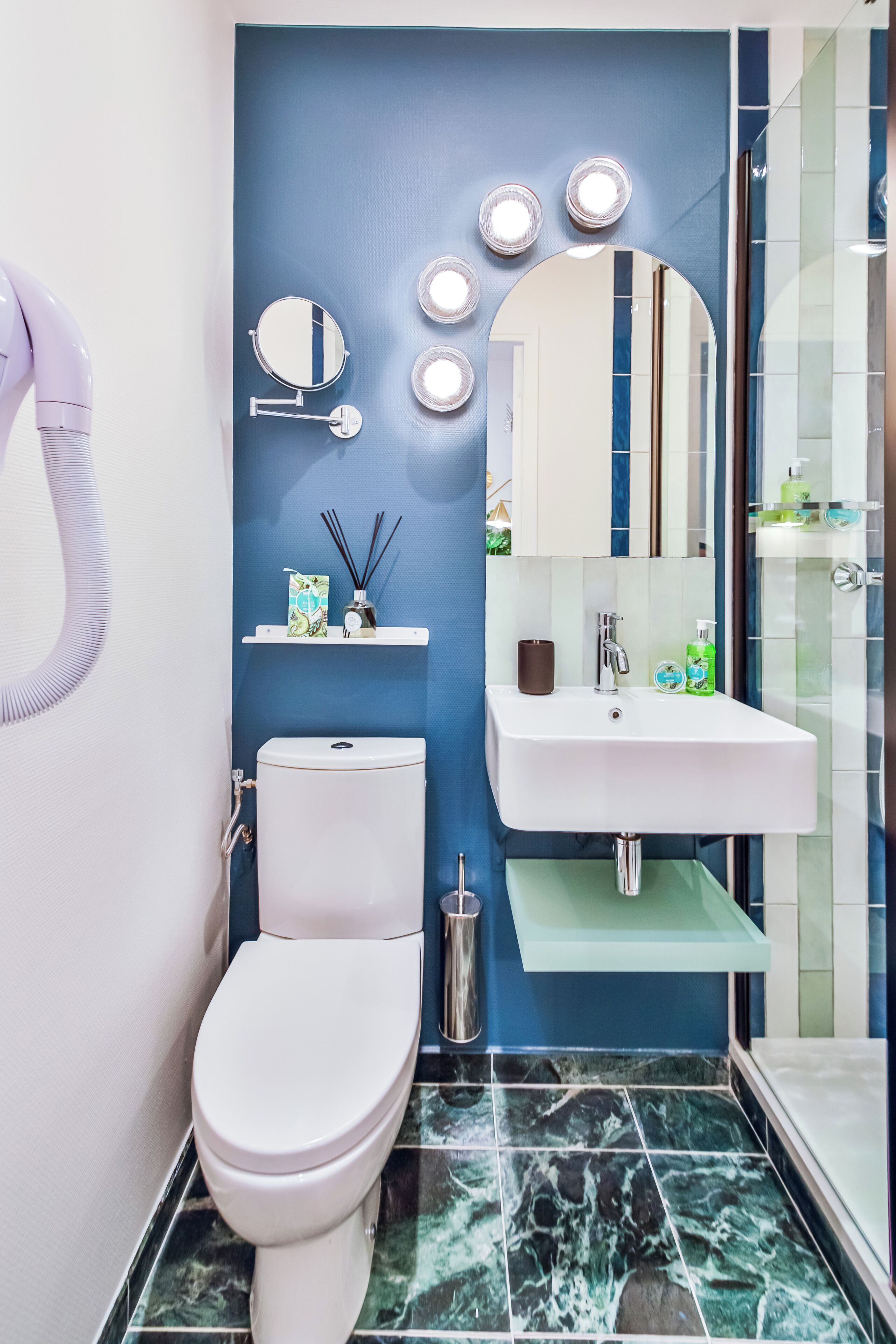 Une salle de bain moderne avec des toilettes blanches, un mur d’accent bleu, un lavabo blanc avec un miroir au-dessus, et un espace douche.
