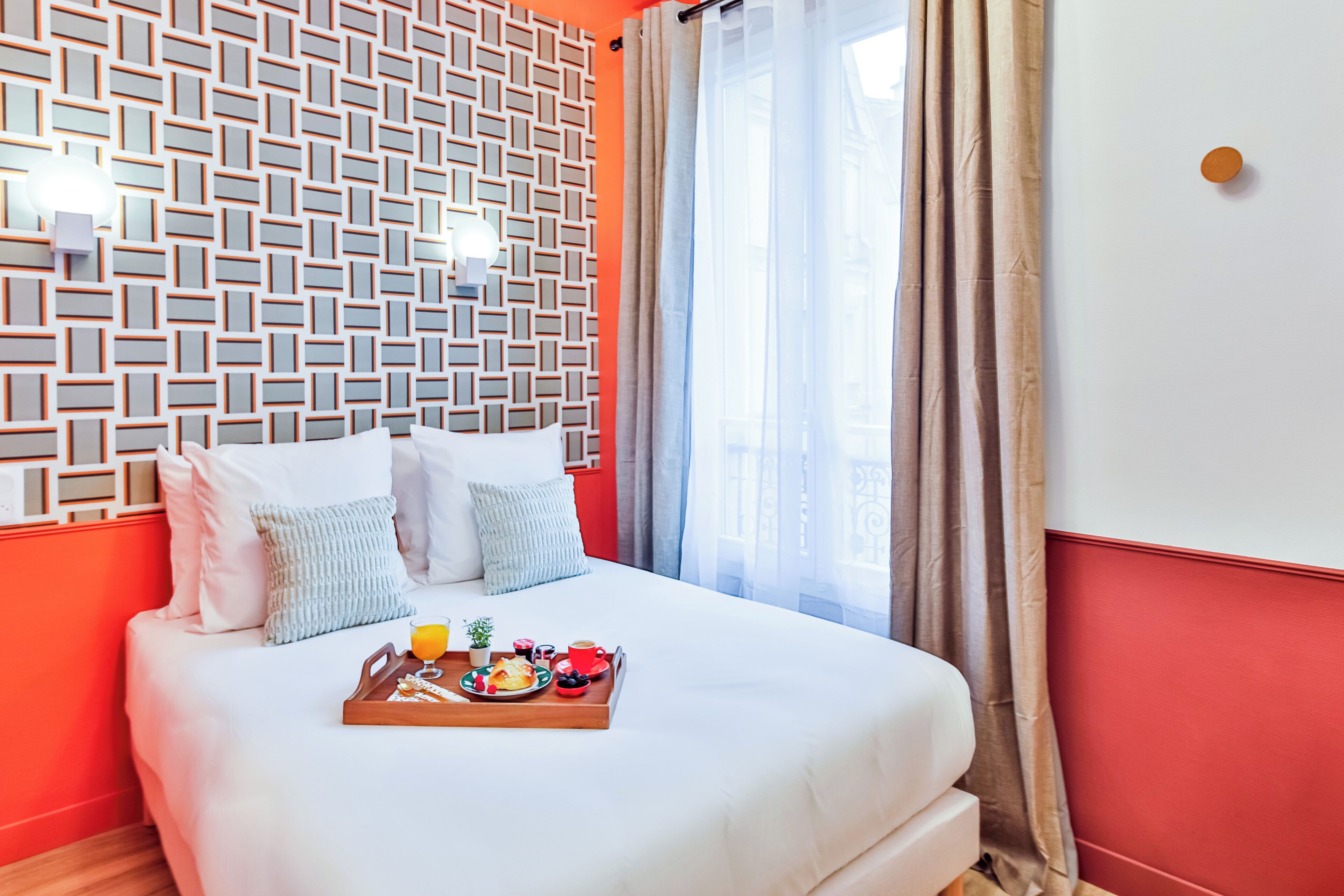 Un lit bien fait, avec un plateau de petit-déjeuner, un mur orné d’un motif et une fenêtre avec des rideaux.