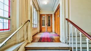 A grand hallway with a wooden staircase, a red carpet, and large windows with a red frame.