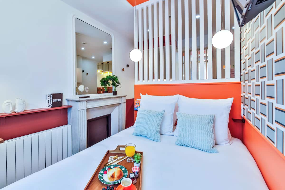 A modern hotel room with a large bed, orange and white decor, a fireplace, and a breakfast tray on the bed.