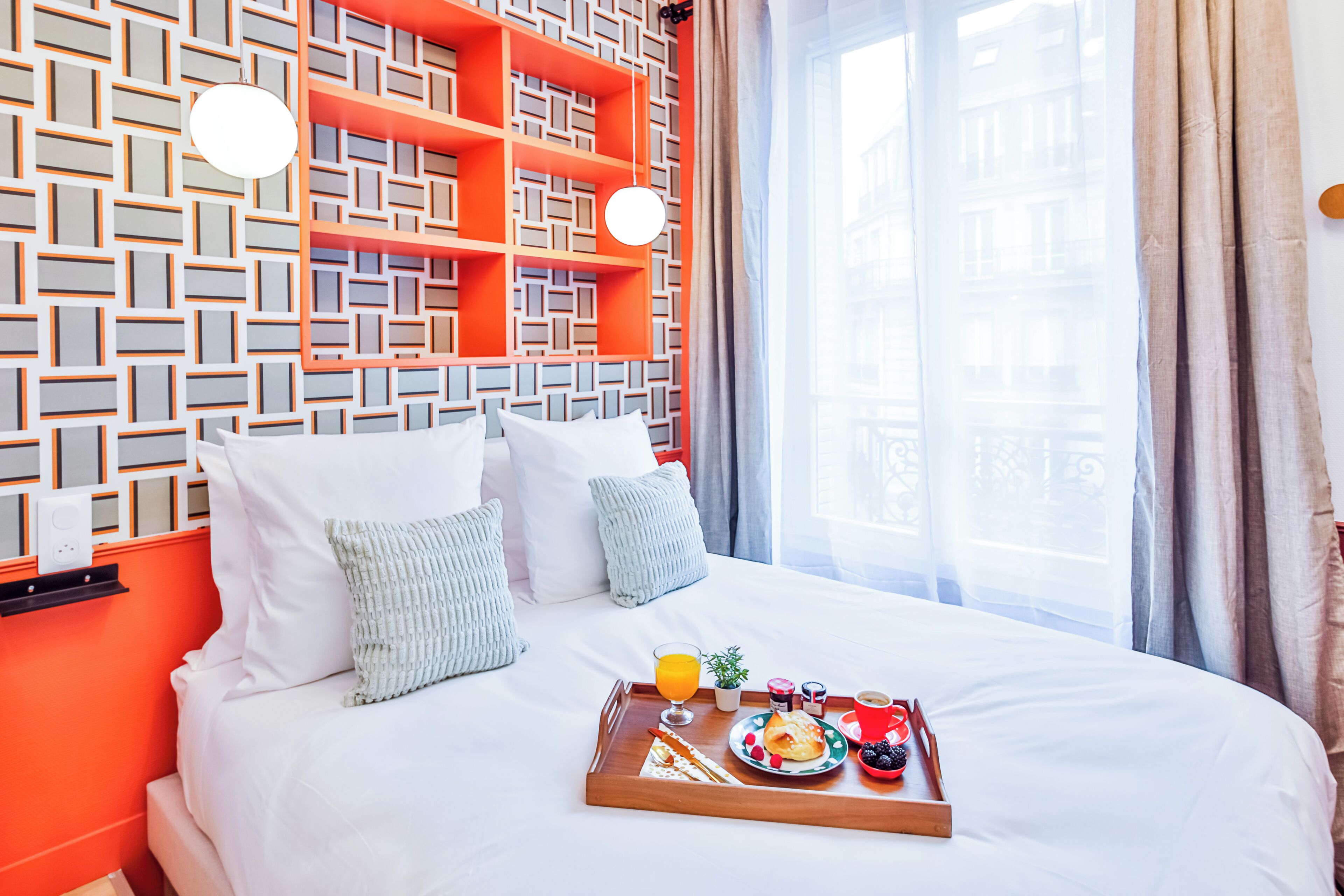 Une chambre d’hôtel dotée d’un décor mural géométrique moderne, d’un lit parfaitement fait avec du linge de lit blanc, et d’un plateau de petit-déjeuner posé sur le lit.