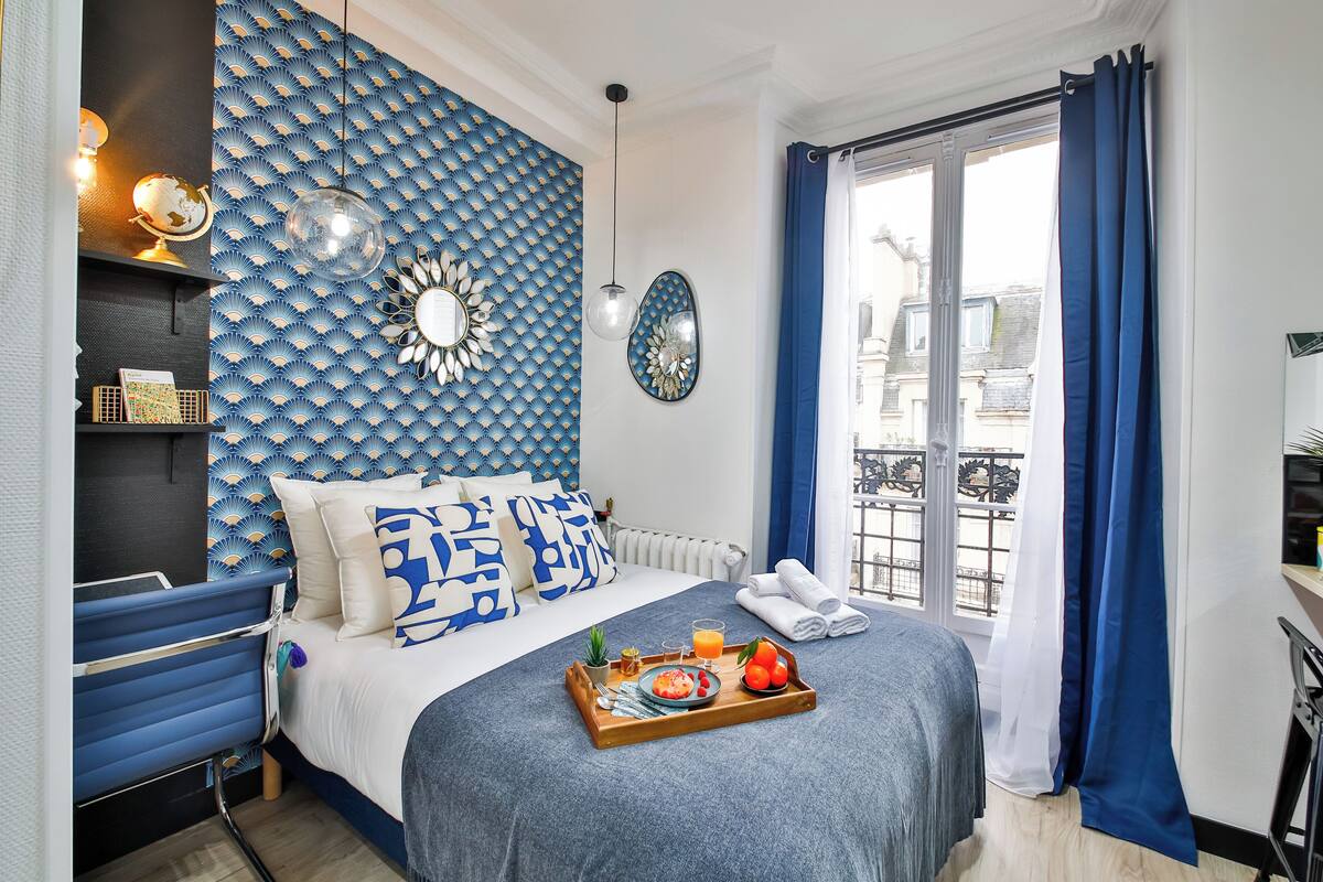 A neatly arranged bedroom with a bed, a bedside table with a globe and lamp, a tray with breakfast items, and a view of the outside through a window with blue curtains.