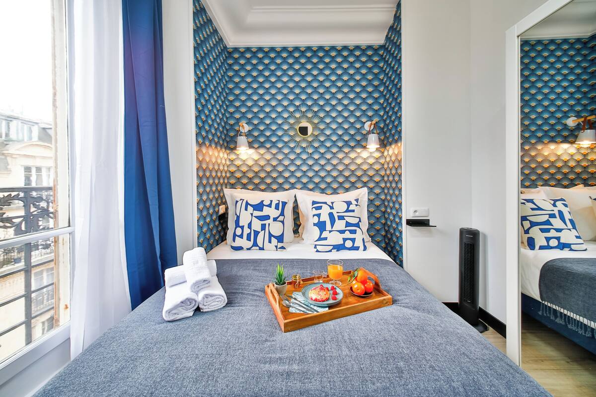 A neatly made bed with a tray of breakfast items, a potted plant, and towels on a window-side bed in a room with patterned blue walls and a view of the city.