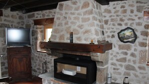 A stone fireplace with a television on a wooden stand to the left, a stone wall with a picture, and a tiled floor. - Stone and wood house (Saint-Pal-de-Chalencon)