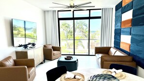 A modern living room with a large TV, leather sofas, a dining table with placemats, and a view of greenery through large windows. - Welcome N/H Villa Paradise in enchanting Reading, Montego Bay with AC, Gym, WiFi (Reading)